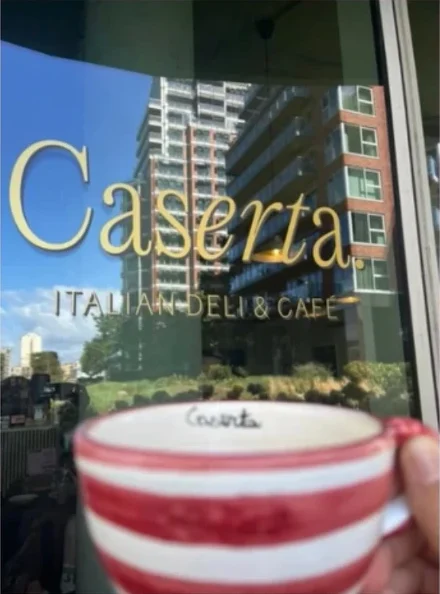 A red-and-white striped Caserta cup held up in front of the café window, lettered “Caserta. Italian deli & café”